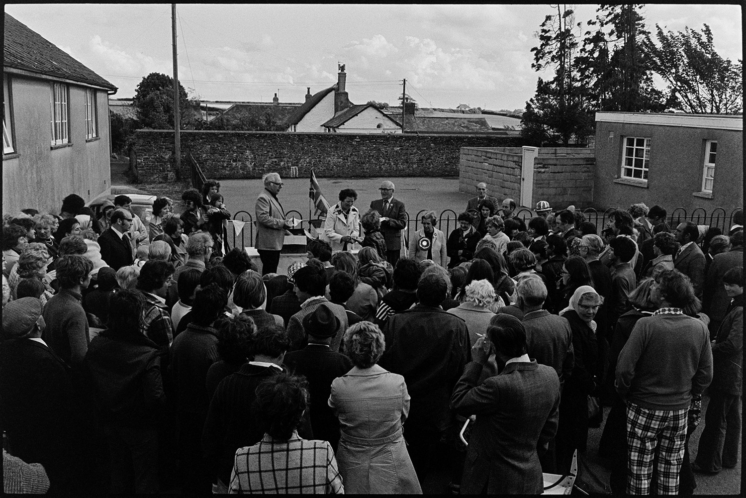 Presentation of mugs in street crowd watching. 
[Mrs Thomas presenting mugs to children in a street in High Bickington to celebrate the Silver Jubilee of Queen Elizabeth II. A crowd is watching the presentation.]
