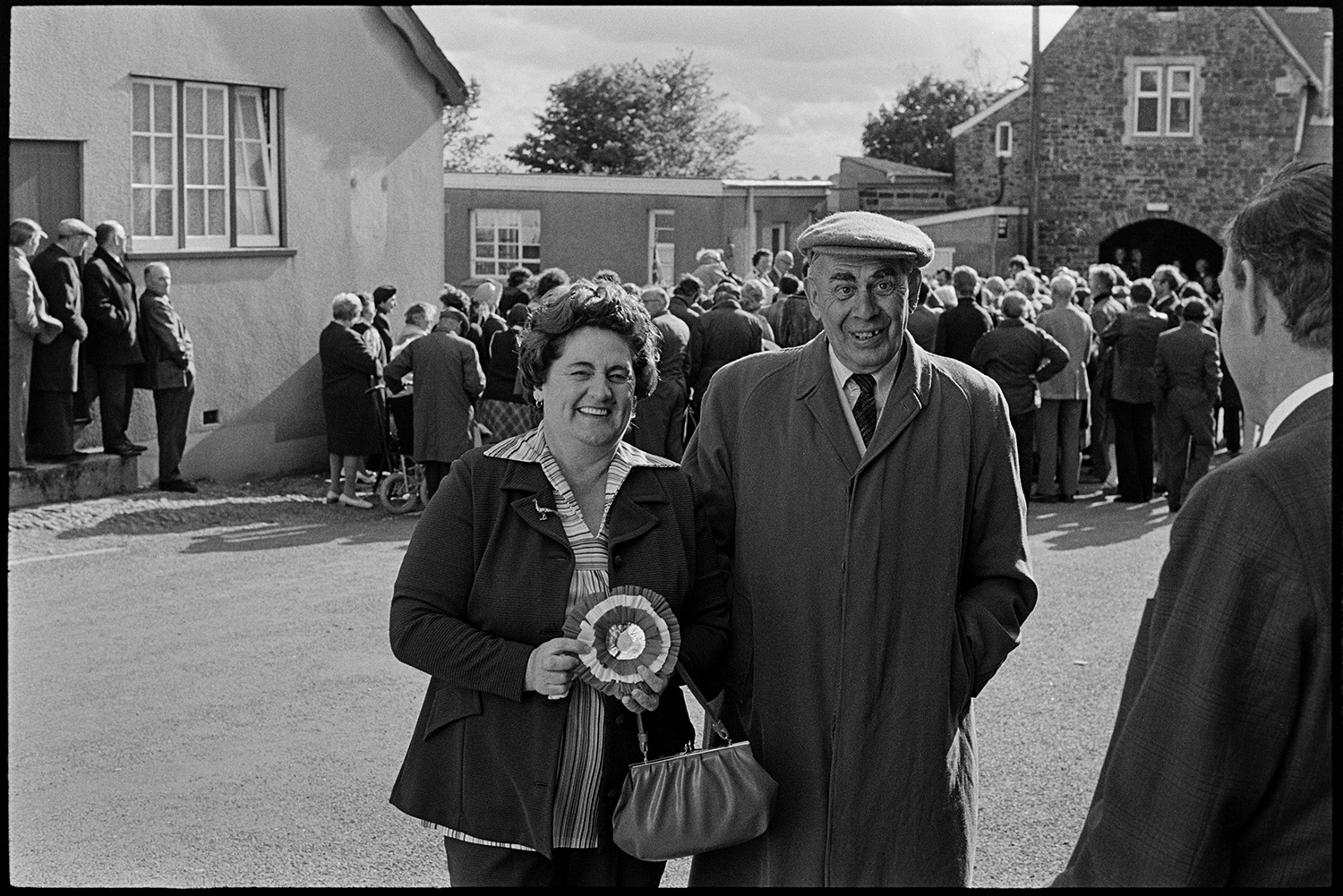 Presentation of mugs in street crowd watching. 
[A man and woman posing for a photograph in a street in High Bickington at the celebrations for the Silver Jubilee of Queen Elizabeth II. The woman is holding a rosette. A crowd in the background are watching a presentation of mugs to children from the village.]
