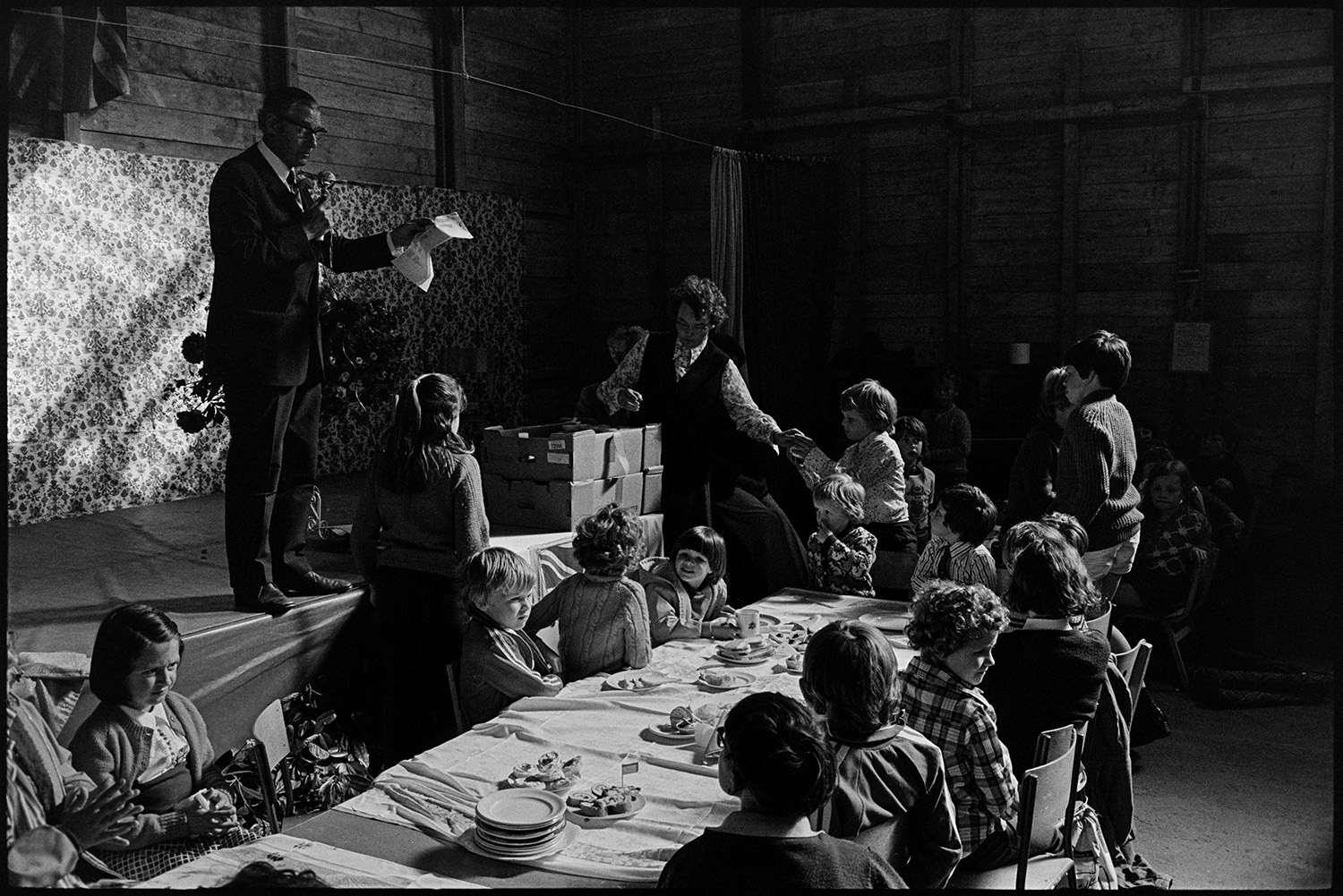 Presentation of Jubilee mugs in warehouse. 
[Children being presented with mugs to celebrate the Silver Jubilee of Queen Elizabeth II in a warehouse or hall in Burrington.]