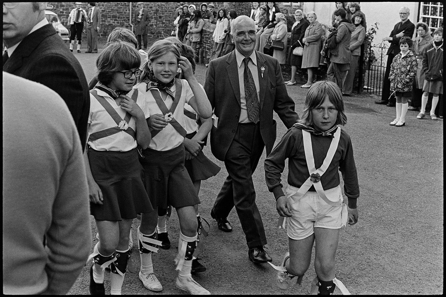 Children Morris dancing in village street. 
[Children Morris Dancers walking through a street in High Bickington with a man after performing in the celebrations for the Silver Jubilee of Queen Elizabeth II. Spectators can be seen in the background.]