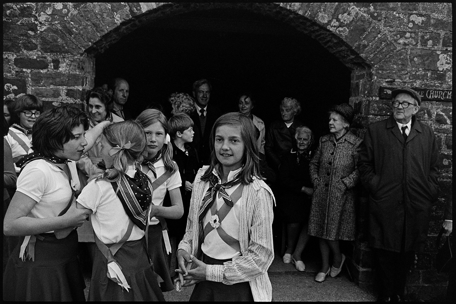 Children Morris dancing in village street. 
[Child Morris Dancers waiting to perform at High Bickington Primary School in the celebrations for the Silver Jubilee of Queen Elizabeth II. Men and women are stood under a stone archway watching them.]