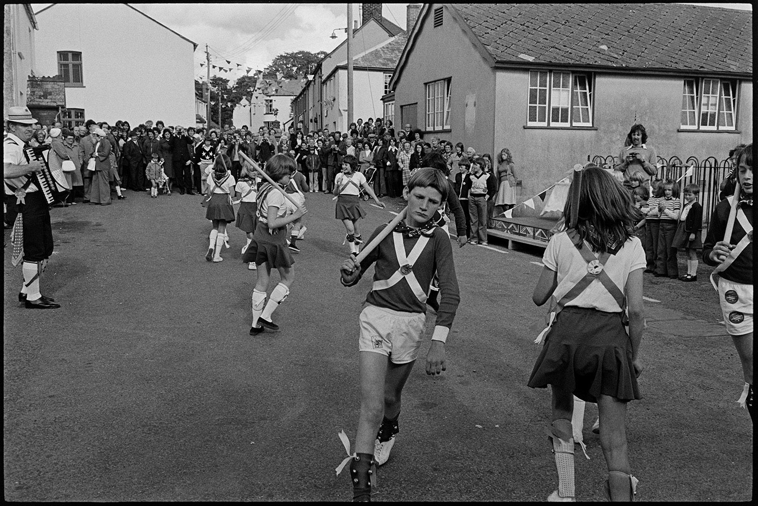 Children Morris dancing in village street. 
[Child Morris Dancers performing in a street in High Bickington as part of the celebrations for the Silver Jubilee of Queen Elizabeth II. A man is playing the accordion for them and a crowd is watching from further up the street. A street in the background is decorated with bunting,]