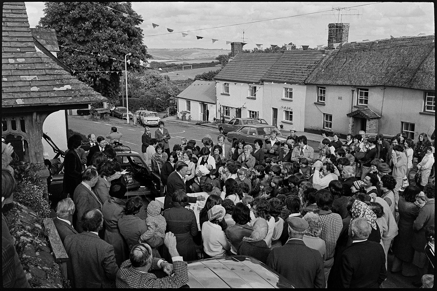 Presentation of Jubilee mugs, crowd in front of mugs set out on table, man presenting them.
[A man presenting mugs to children in Atherington, to celebrate the Silver Jubilee of Queen Elizabeth II. A crowd of men and women are watching. The village square is decorated with bunting, with thatched cottages in the background.]