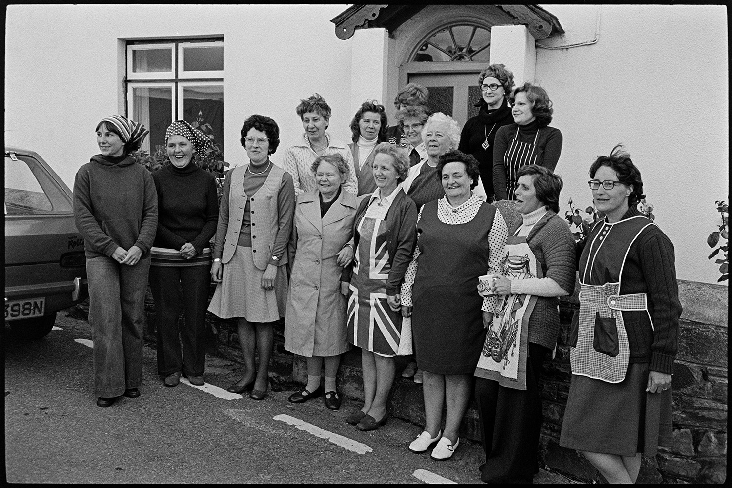 Group of women organisers who worked, photographer taking their photograph. 
[A group of women posing for a photographer, Chris Hammond, outside a house in Atherington, after preparing a lunch to celebrate the Silver Jubilee of Queen Elizabeth II. One of the women is wearing a Union Jack apron.]