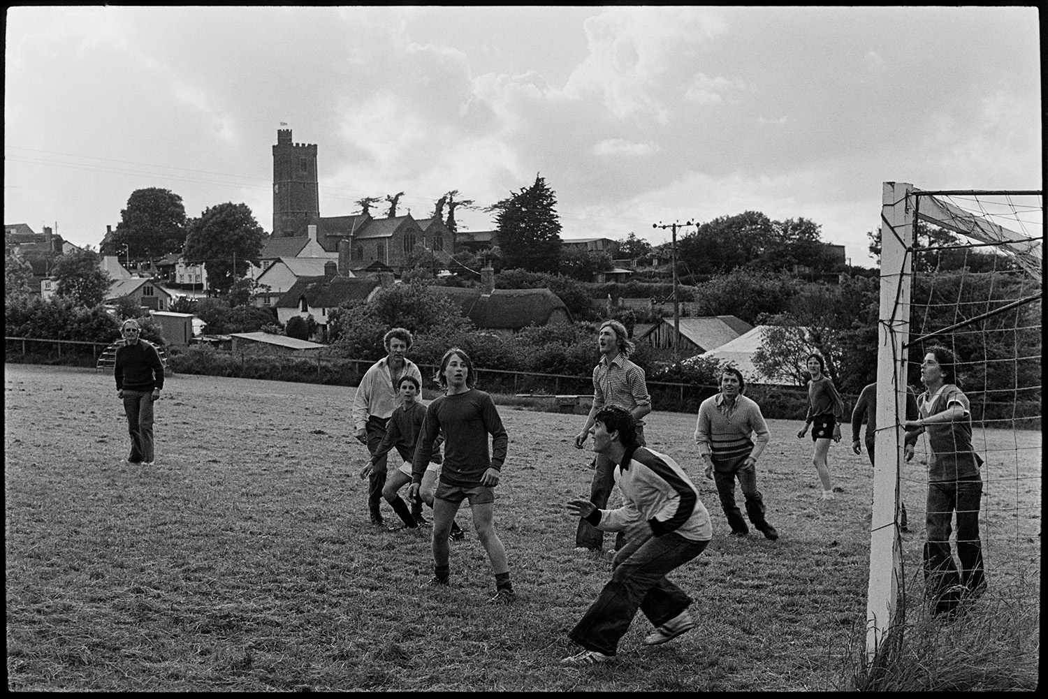 Jubilee football match.
[Young men playing football during the Silver Jubilee celebrations of Queen Elizabeth II in Atherington. Thatched cottages and Atherington church tower can be seen in the background.]