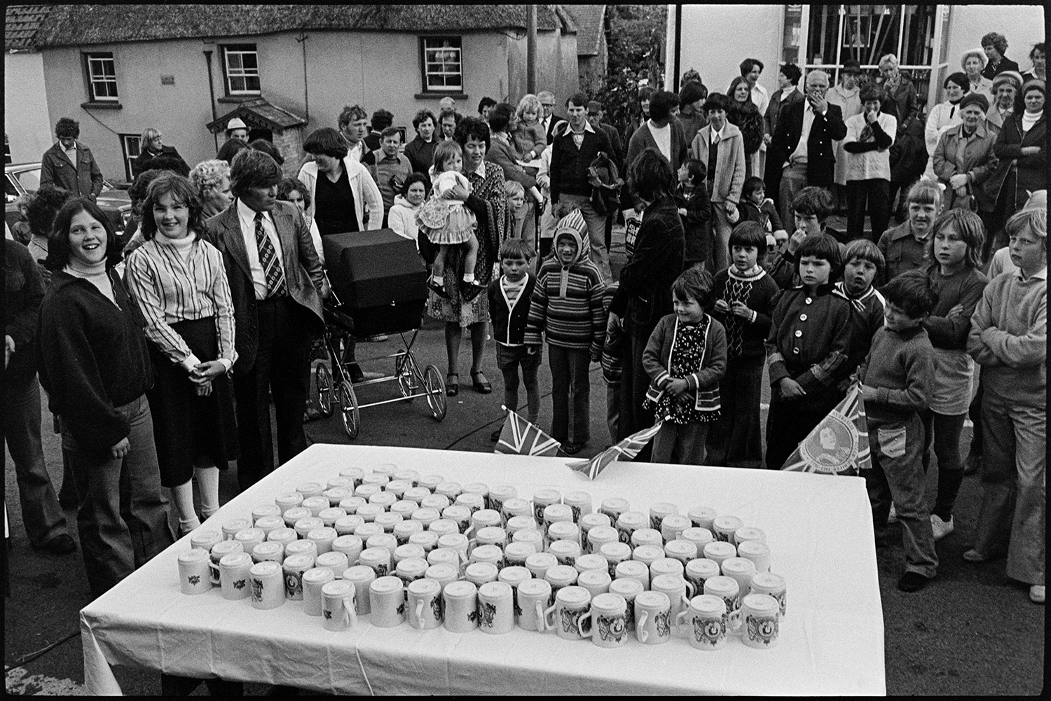 Presentation of Jubilee mugs, crowd in front of mugs set out on table, man presenting them.
[A group of men, women and children standing in Atherington village square around a table with presentation mugs to celebrate the Silver Jubilee of Queen Elizabeth II.]