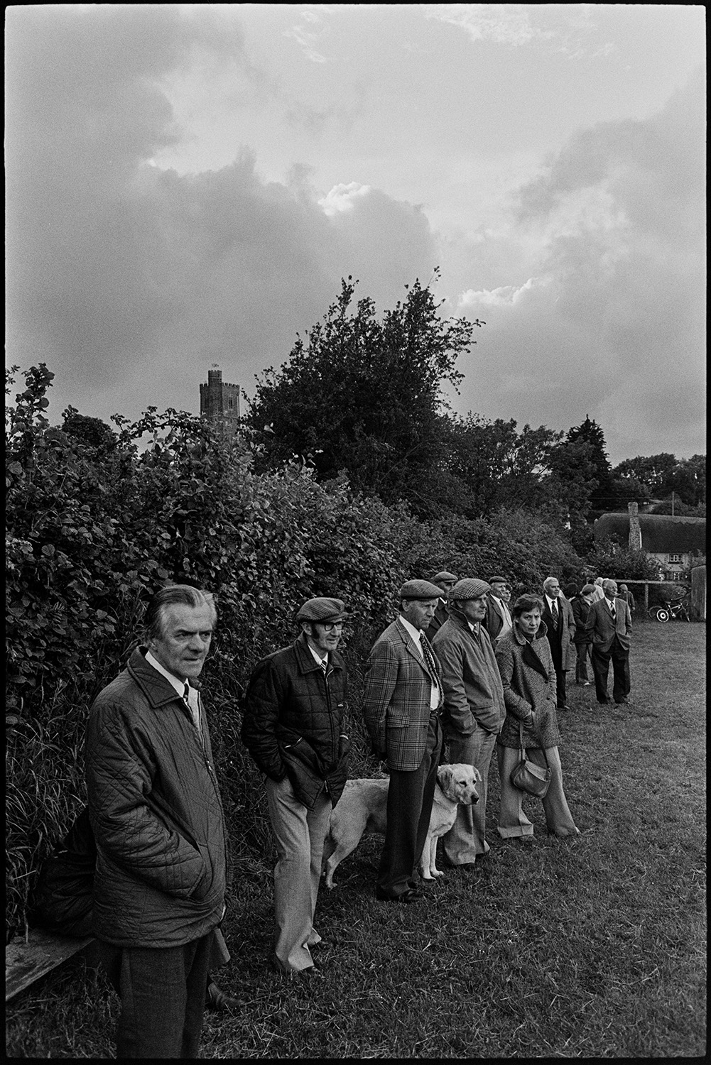 Jubilee football match with spectators. 
[Spectators at village football match in Atherington to celebrate  the Silver Jubilee of Queen Elizabeth II. One of the spectators has a dog and Atherington Church tower is visible in the background.]