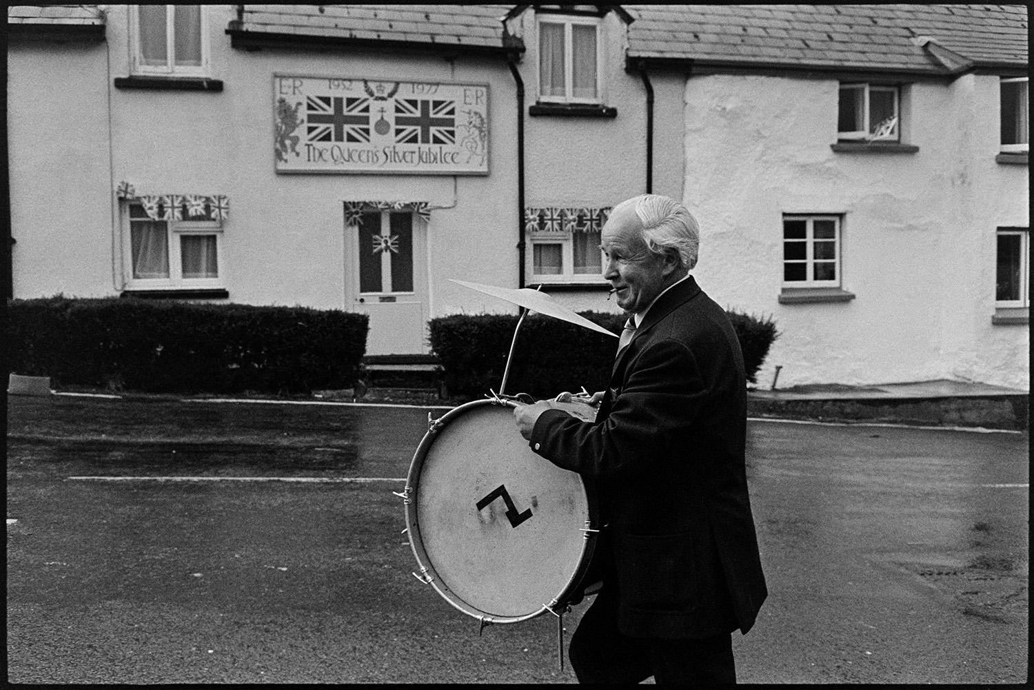 Bandsmen and choir ladies in street. 
[A man carrying a drum and cymbal in a street in Atherington. He is passing a cottage with a  sign above the door and flags celebrating the Silver Jubilee of Queen Elizabeth II.]
