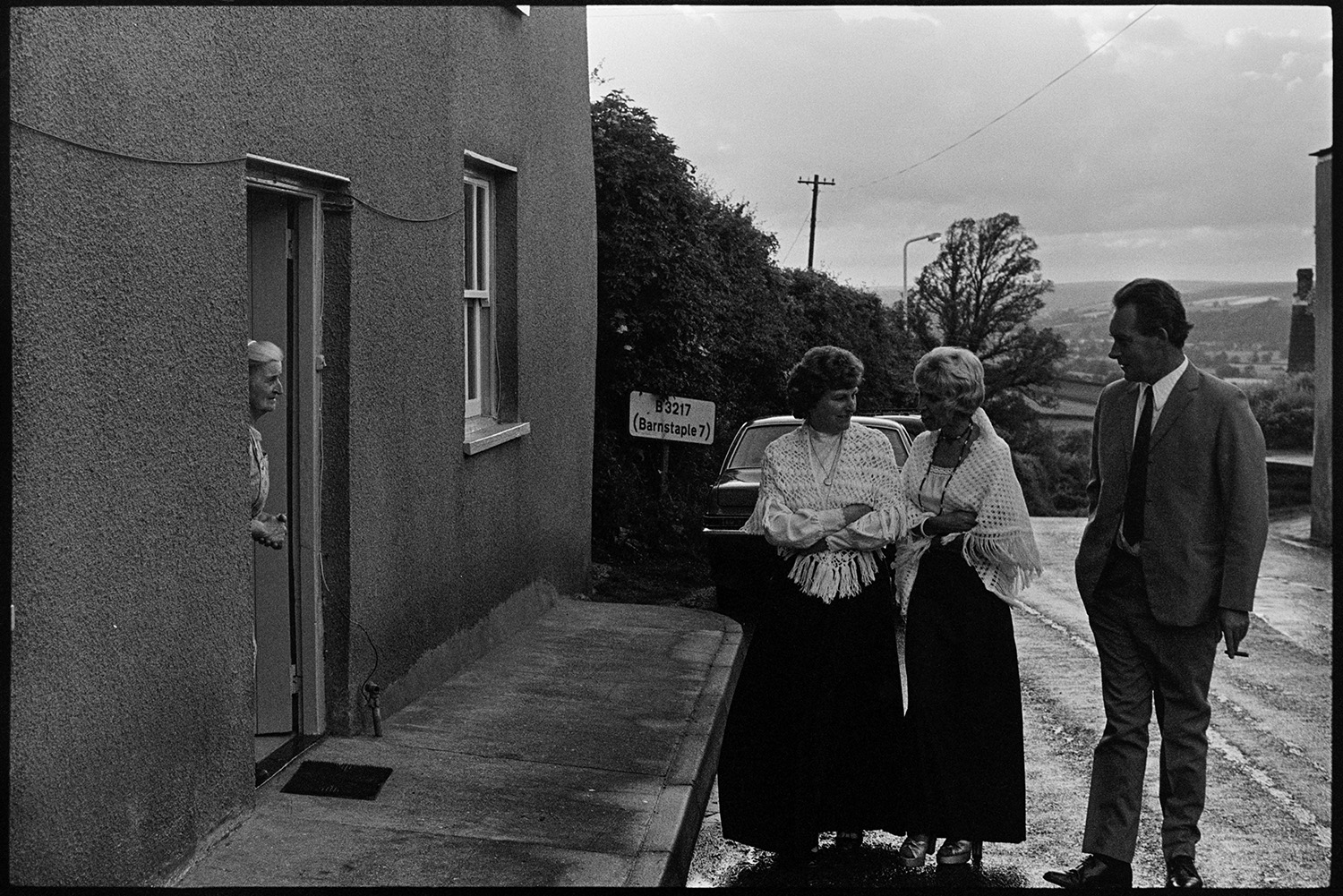 Bandsmen and choir ladies in street. 
[Two women wearing shawls, and a man wearing a suit, standing in a street in Atherington talking to Norah Maynard looking out of her doorway, during the celebrations for the Silver Jubilee of Queen Elizabeth II. The house was formerly the White Hart Inn.]