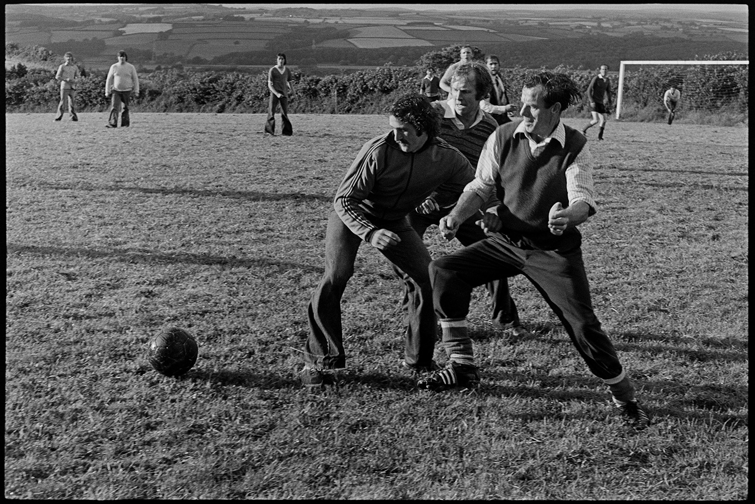 Jubilee football match with spectators. 
[Men playing a football match in Atherington to celebrate the Silver Jubilee of Queen Elizabeth II. Three men are tackling each other for the leather ball. A rural landscape of fields can be seen in the background.]