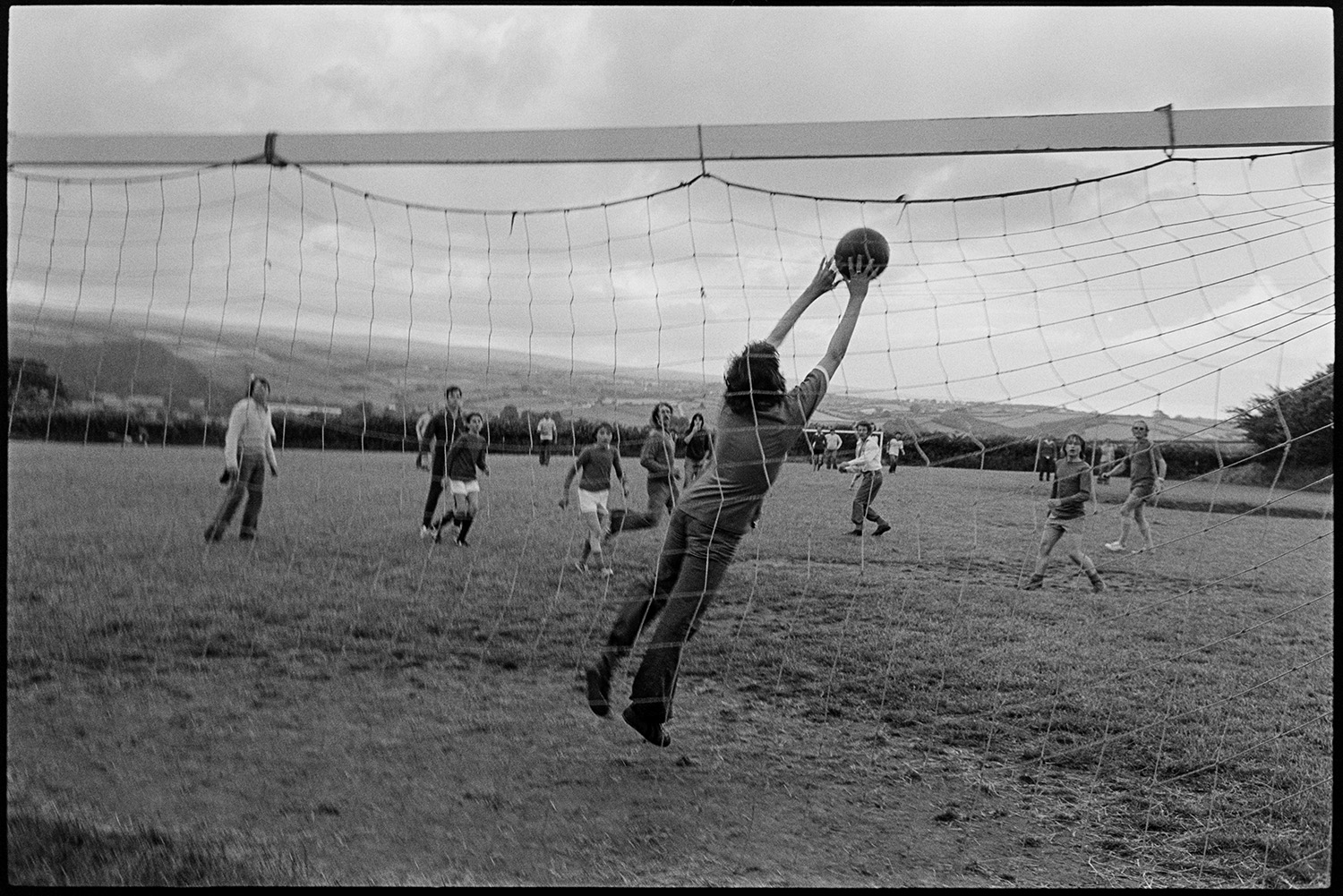 Jubilee football match with spectators. 
[Young men playing football in Atherington during the Silver Jubilee celebrations of Queen Elizabeth II. The goal keeper is jumping to save a goal. A rural landscape and some spectators are visible in the background.]