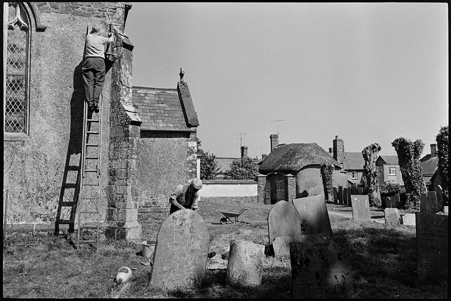 Vicar and helpers repairing church.
[Two men doing repair work at Kings Nympton church. One man is up a ladder repointing stonework while another man is working behind a gravestone, possibly mixing mortar. Pollarded trees can be seen in the churchyard and thatched cottages and houses are visible in the background.]