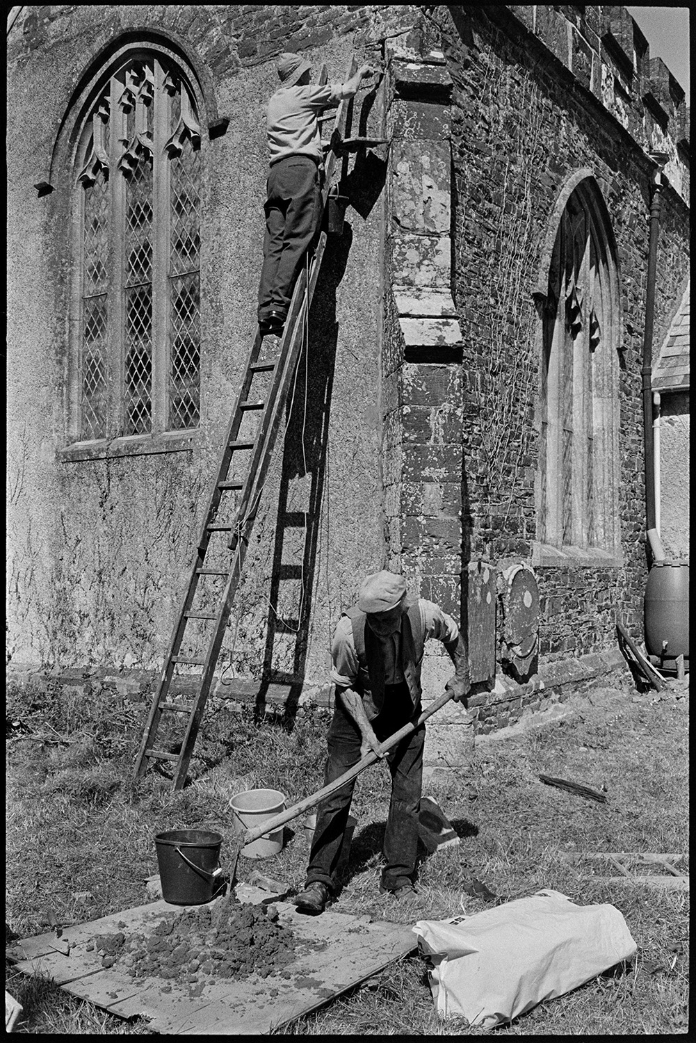 Vicar and helpers repairing church.
[A man up a wooden ladder repointing the stonework at Kings Nympton Church. Church windows and stone buttress are visible. Another man is mixing mortar with a spade, on wood planks in the churchyard.]