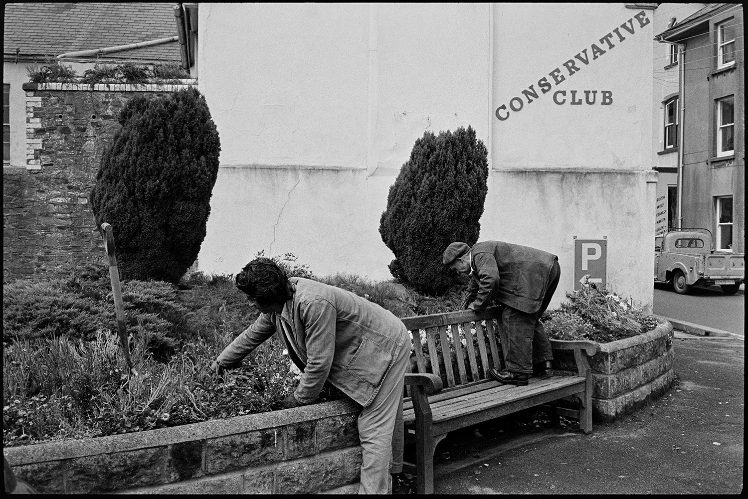 Street scenes with men tending flowers, motor bikes. 
[Two council workmen weeding flower beds in Torrington town centre. A fork is in the flower bed. One man is standing on a wooden bench to weed the flower bed. A Conservative Club and parking sign are on the wall behind the flower bed.]