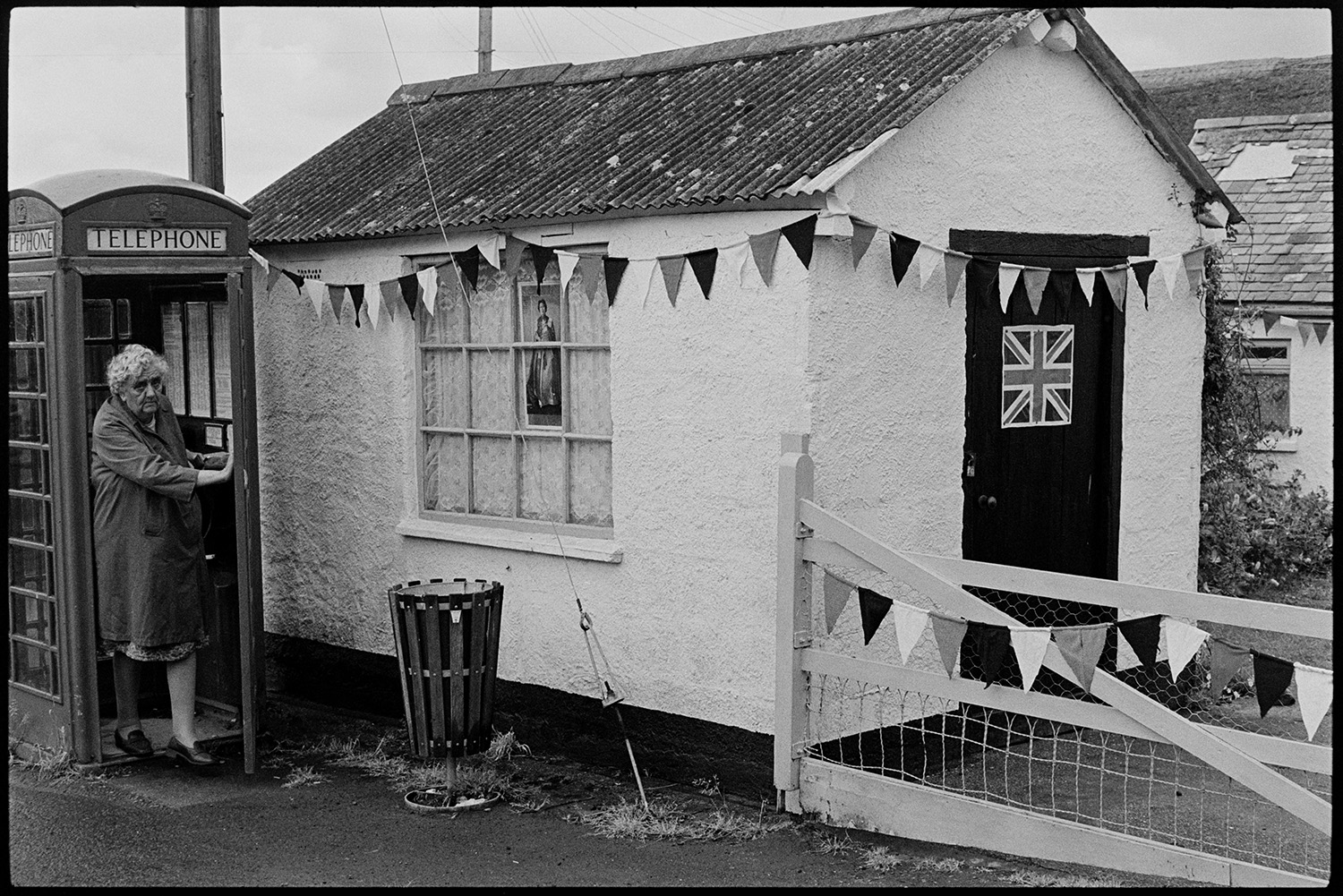 Decorated shed and telephone kiosk at Jubilee time.
[A woman coming out of a telephone box in Kings Nympton, next to a small building with a corrugated iron roof. The building is decorated with a union jack flag on the door, a photograph of the Queen in the window, and bunting for  the Silver Jubilee of Queen Elizabeth II.]