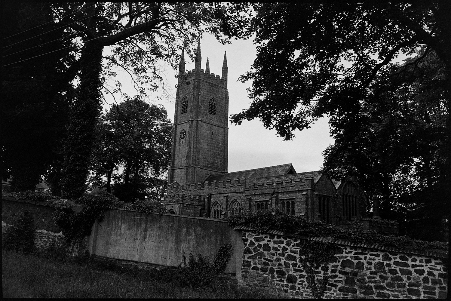 View of church with tower and also church entrance with wooden gate.
[Sampford Courtenay Church tower showing the church clock. A stone wall with tiles and trees surround the church.]