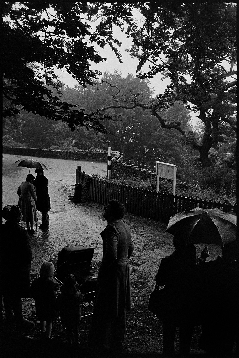 Tourists in the rain at entrance to Clovelly, umbrellas.
[Tourists in the rain, holding umbrellas, about to walk down the wooded valley at Clovelly. One woman has a pram and two small children.]