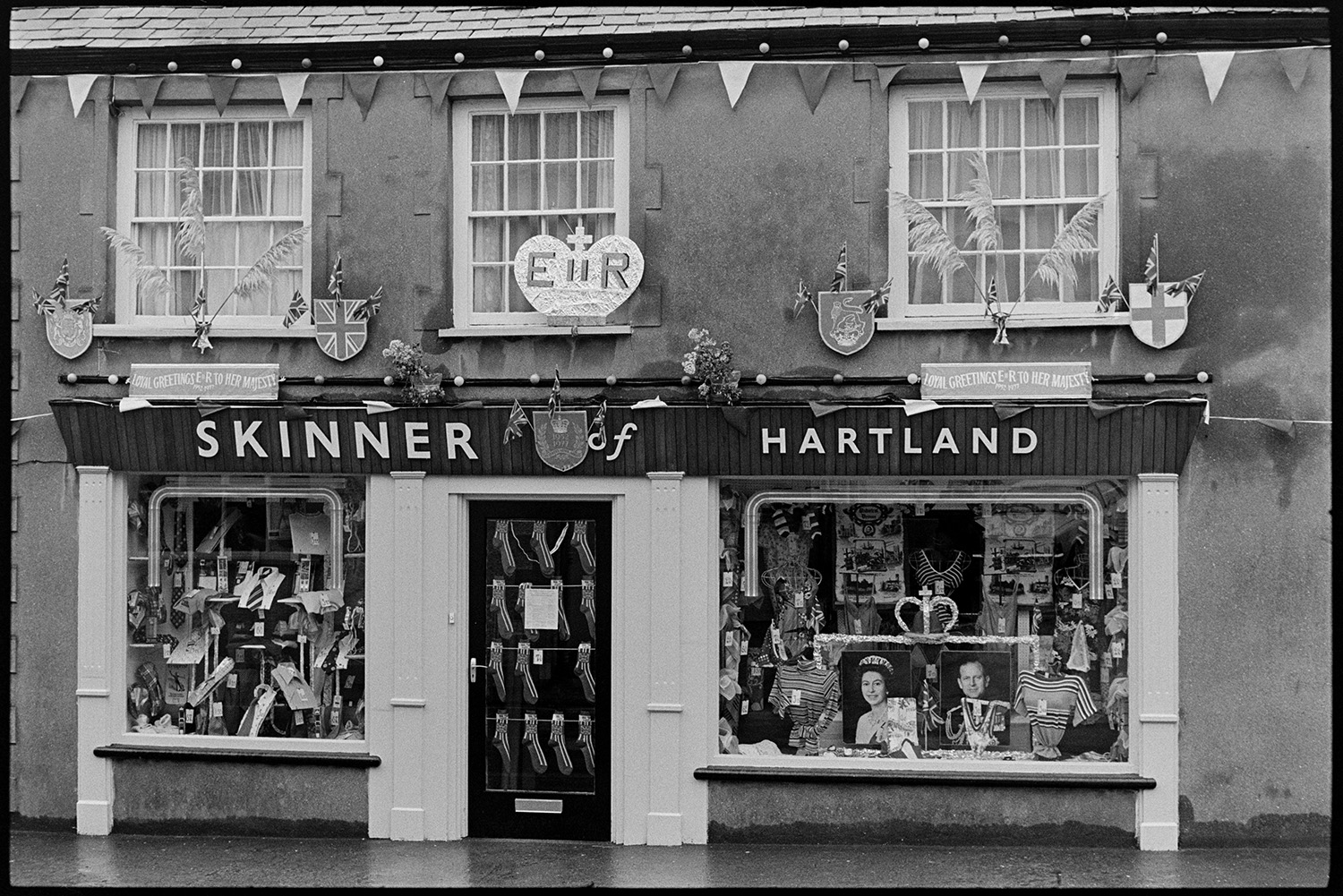 Clothes shop decorated for Jubilee, socks hanging in door.
[The shop front window of the Skinner of Hartland clothes shop in Hartland. It is decorated with Union Jack flags, crown motifs and pictures of the Queen to celebrate the Silver Jubilee of Queen Elizabeth II. Socks with a Union jack design are hung up on the front door.]