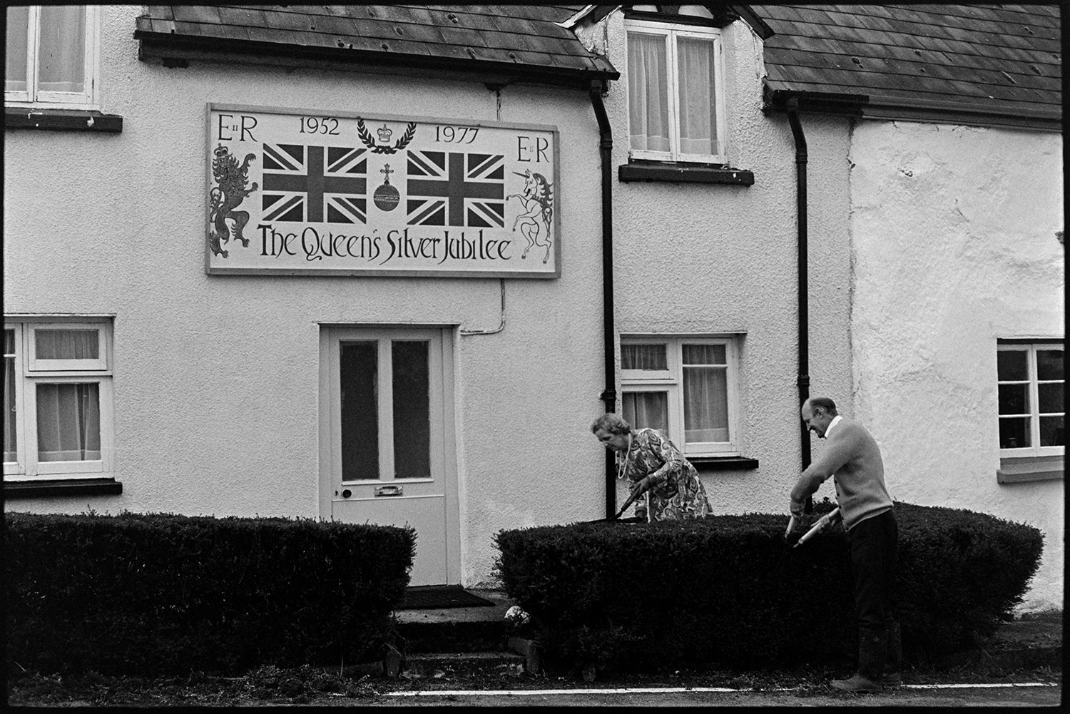 House with large painting of flags etc for Jubilee artist clipping hedge.
[Geoff Siviter and Barbara Siviter trimming the hedge in front of their house in Atherington. The house has a large painted sign above the front door to celebrate the Silver Jubilee of Queen Elizabeth II.]