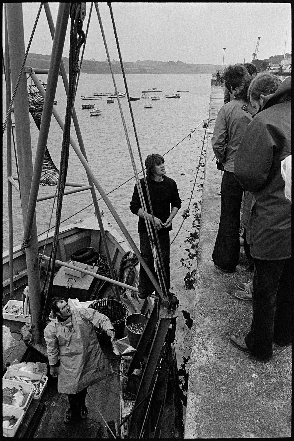 Fishermen selling fish from boat at quayside. 
[Two fishermen selling their catch of fish from their fishing boat to customers on the quayside at Appledore. Other boats can be seen in the estuary in the background.]