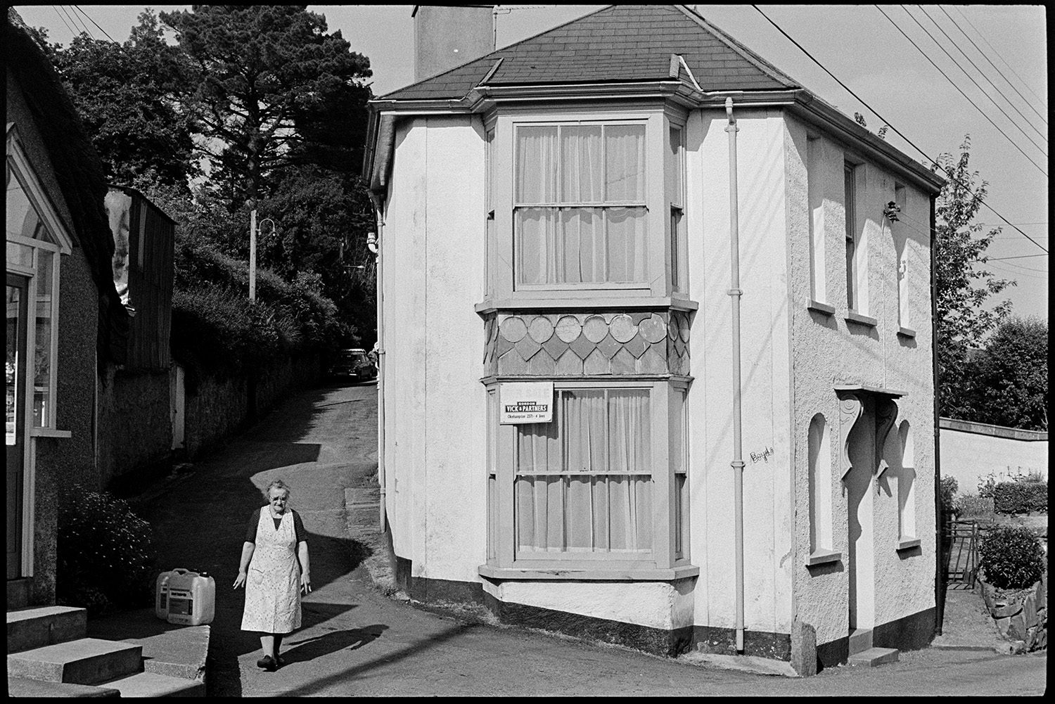 Cottage with decorative slate work. 
[A cottage on the corner of Park Road, Hatherleigh. There is decorative slate work between the bay windows. A woman is walking past the cottage and two containers can be seen further up the road.]