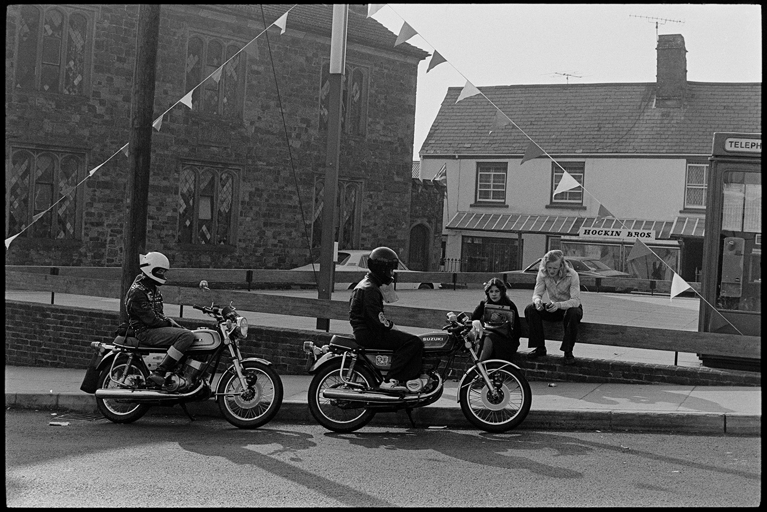Street scenes, motorbikes. 
[Two people on motorcycles in Market Street, Hatherleigh. Two other teenagers are sat on a wall in the square. A telephone box and Hockin Bros shop front are visible in the background.]