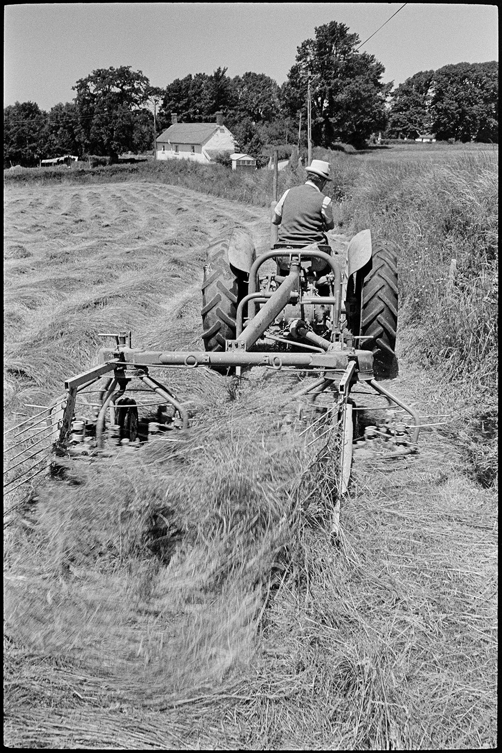 Farmer whisking hay.
[John Ward turning hay with a whisk and tractor in a field at Parsonage, Iddesleigh. A house is visible in the background.]