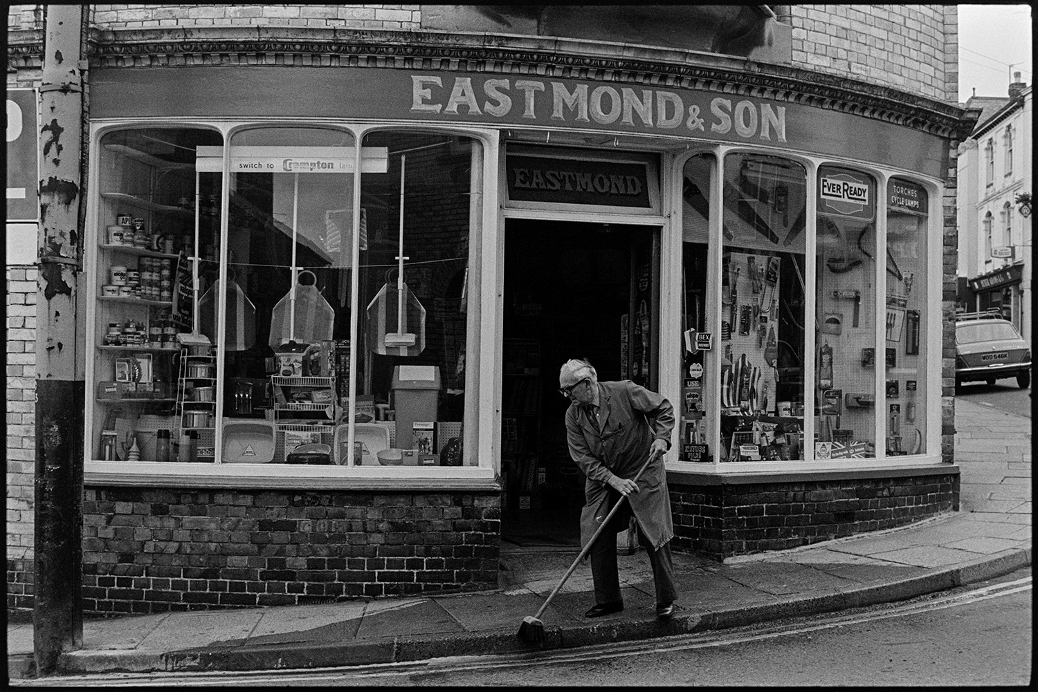 Front of Ironmongers shop, proprietor sweeping pavement. 
[The shop front of Eastmond & Son Ironmongers shop in Torrington. Mr Eastmond is using a brush to sweep the pavement outside the shop. Various goods are on display in the shop window, including saws. washing up bowls and saucepans.]