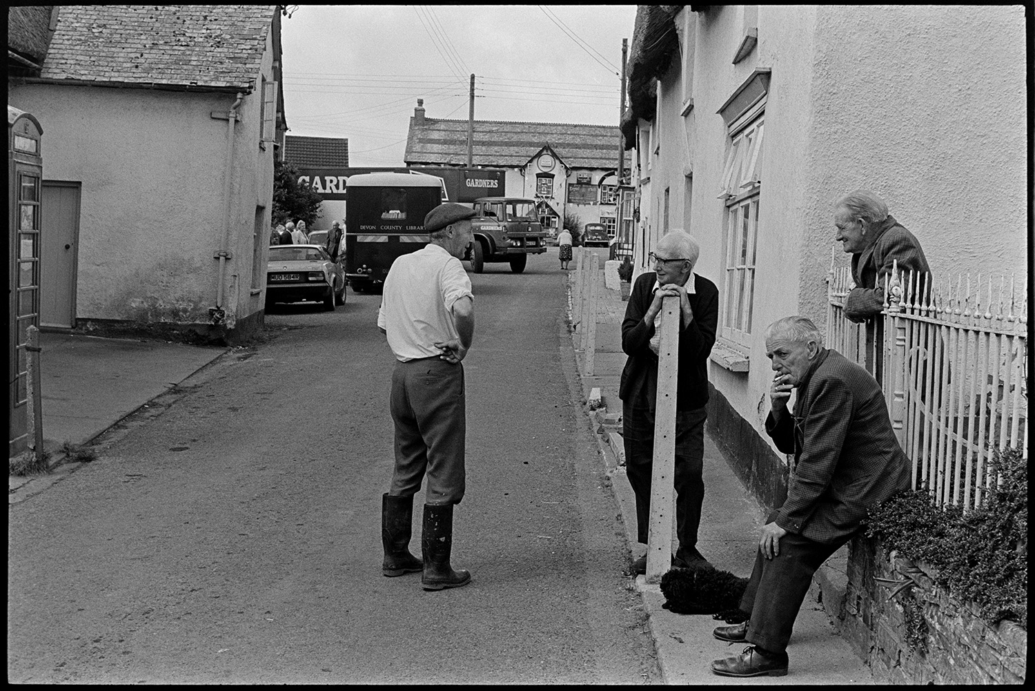 Men chatting in street. 
[A group of men talking in Fore Street, Dolton. One man is leaning on a step ladder, another on railings and one man is sat on a wall, smoking a cigarette. In the background a library van, lorry and Royal Oak pub are visible.]