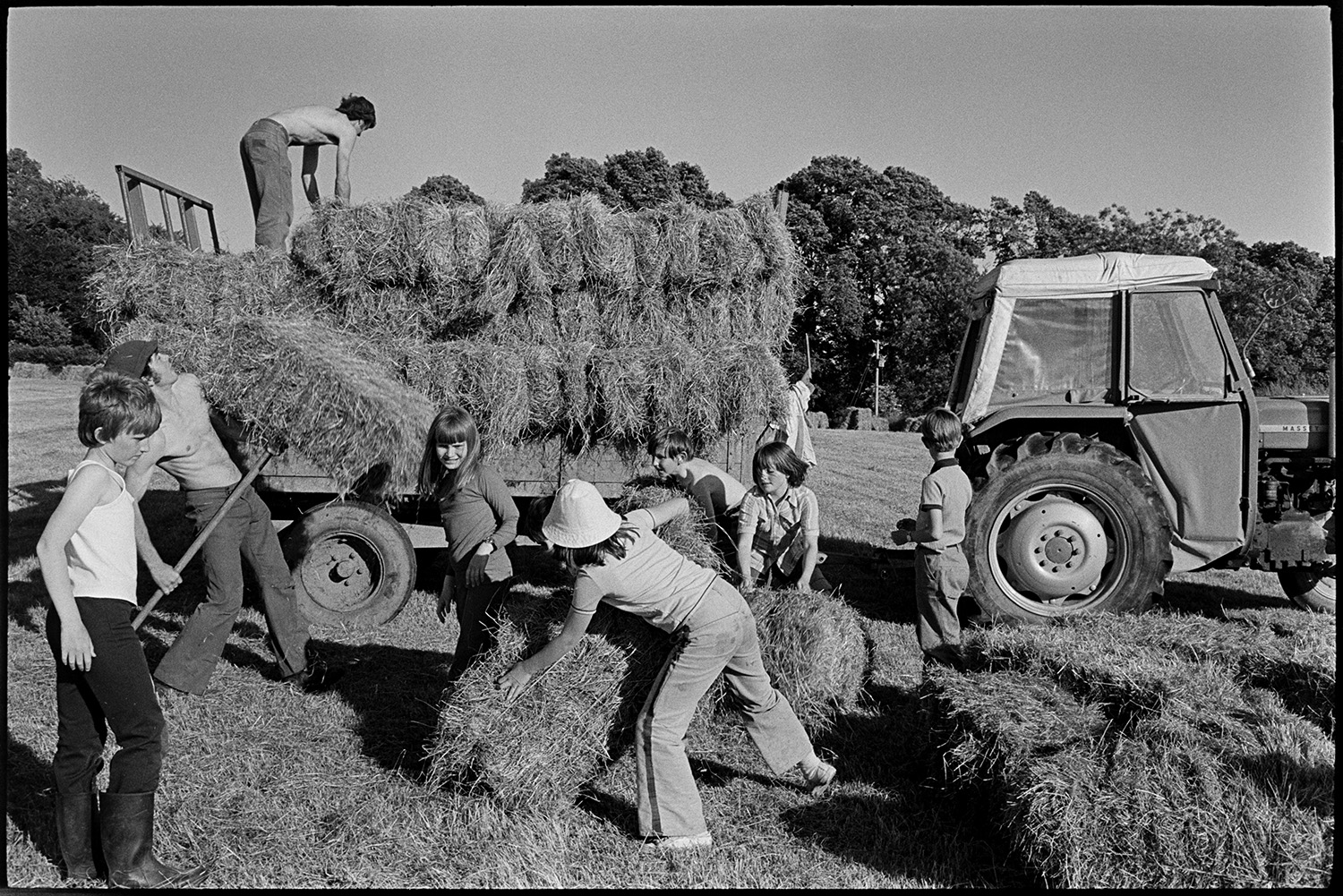 Baling hay with tractor. 
[Children helping Graham Ward and David Ward moving hay bales and load them onto a trailer in a field at Parsonage, Iddesleigh, possibly as part of the Farms for City Children initiative. One of the men is moving a hay bale with a pitchfork.]