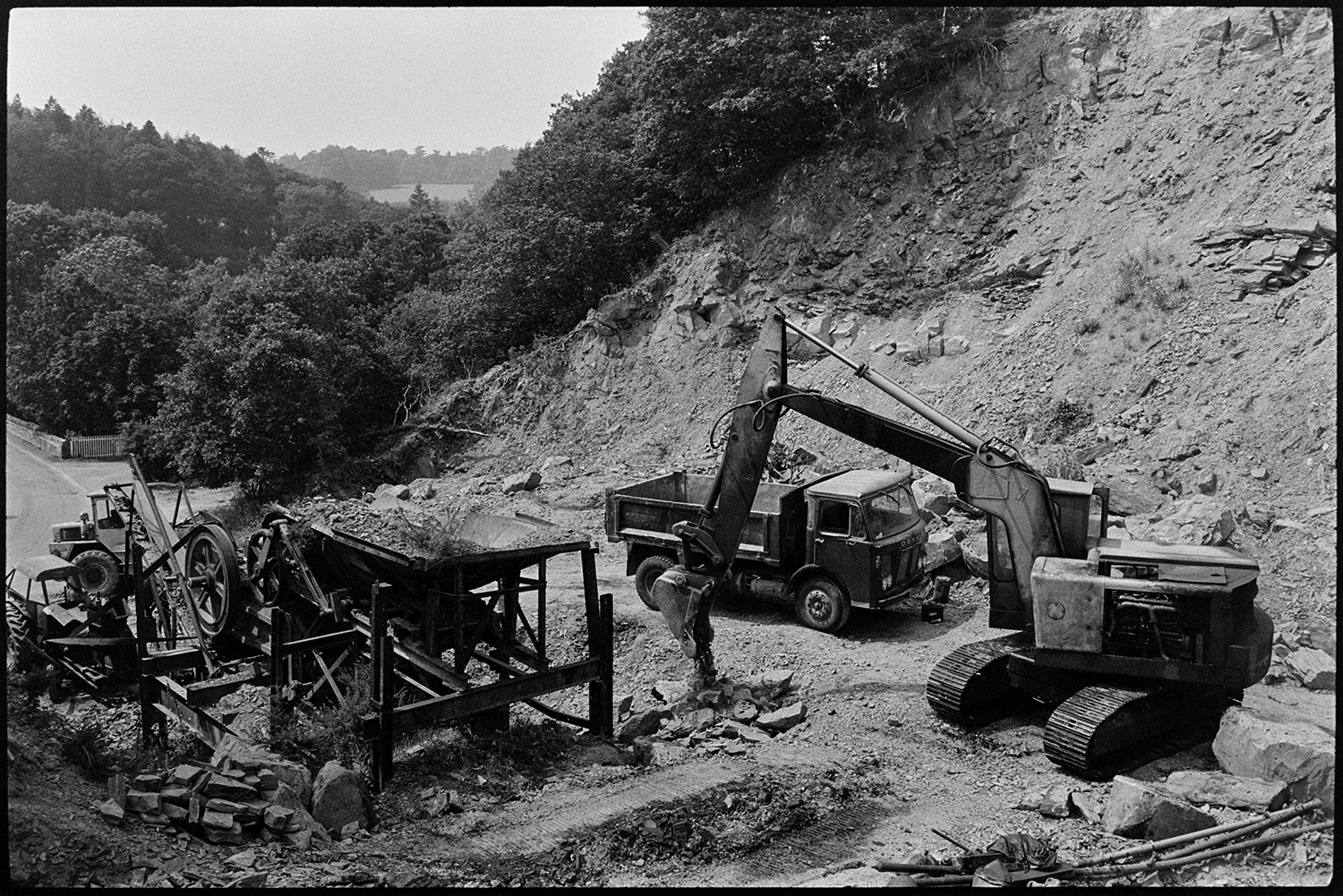 Excavator, crusher and abandoned bulldozer in quarry. Man at controls of excavator. 
[A digger, truck, crusher and other machinery at New Bridge Quarry, Dolton. Woodland can be seen in the background.]