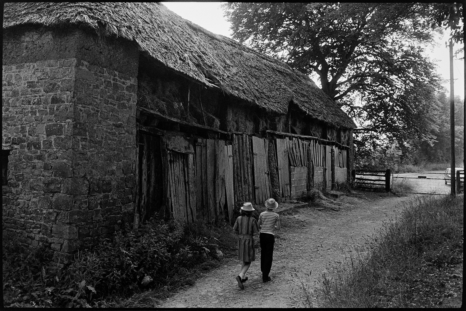 Old thatched barn, extremely interesting. 
[A boy and girl walking past a stone and thatch barn at Deckport, Hatherleigh. Hay bales are being stored in the barn. The bottom part of the barn is covered with sheets of wood and corrugated iron.]