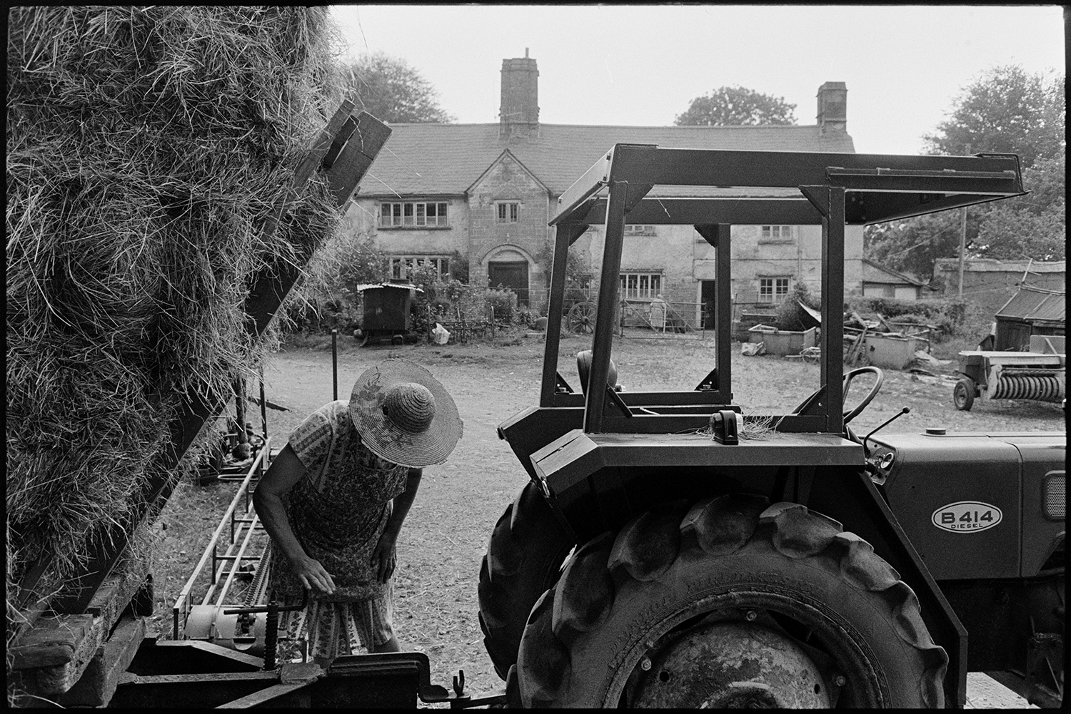 Farming family setting off to bring in the hay on trailer. 
[Mrs Oke checking the connection between a tractor and trailer in the farmyard at Deckport. Hatherleigh. The trailer is full of hay bales and the farmhouse can be seen in the background.]