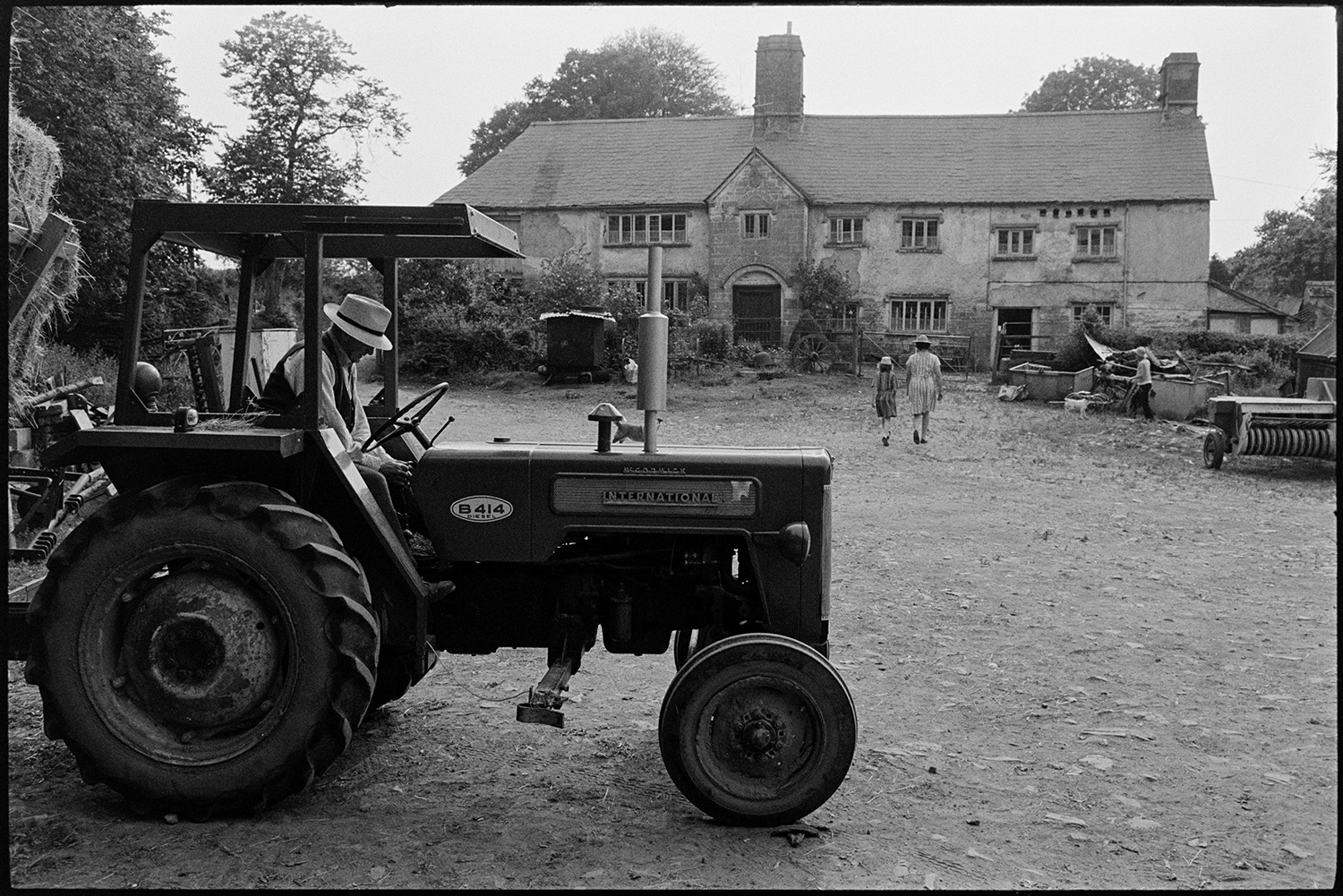 Farming family setting off to bring in the hay on trailer. 
[Mr Oke driving a tractor and trailer with hay bales in the farmyard at Deckport, Hatherleigh. Mrs Oke and her daughter can be seen walking back toward the farmhouse.]