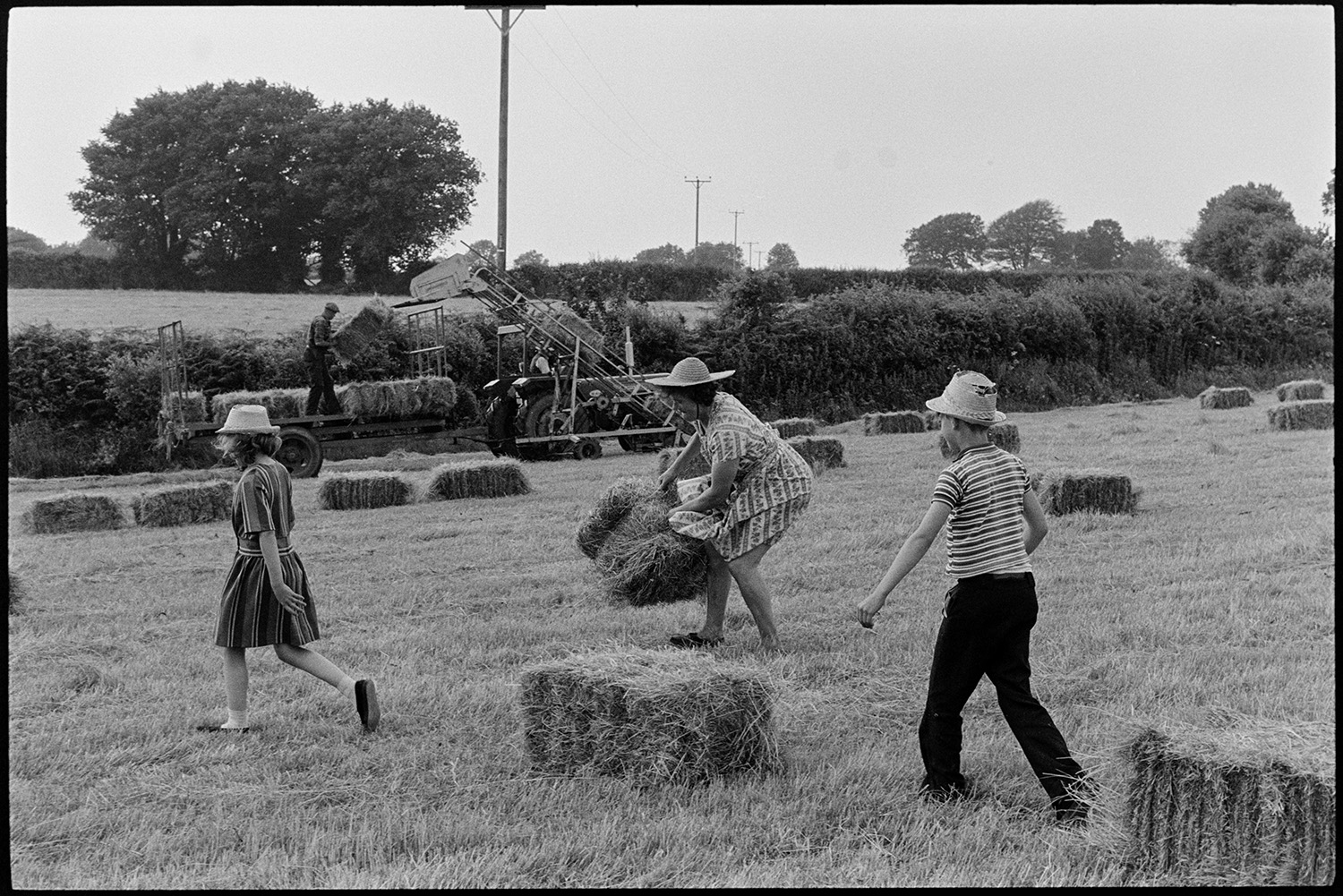 Woman, twin children haymaking. 
[Mrs Oke and her twin children moving hay bales in a field at Deckport, Hatherleigh. Mr Oke is loading the bales onto a tractor and trailer in the background, using an elevator. Mrs Oke is wearing a dress and straw hat.]