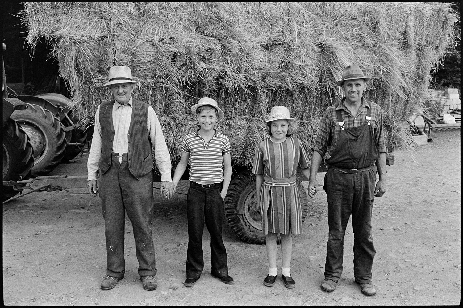 Various groups and shots of farming family. 
[Mr Oke, his daughter and son, who are twins, and another man (right to left) standing in front of a trailer loaded with hay bales in the farmyard at Deckport, Hatherleigh.]