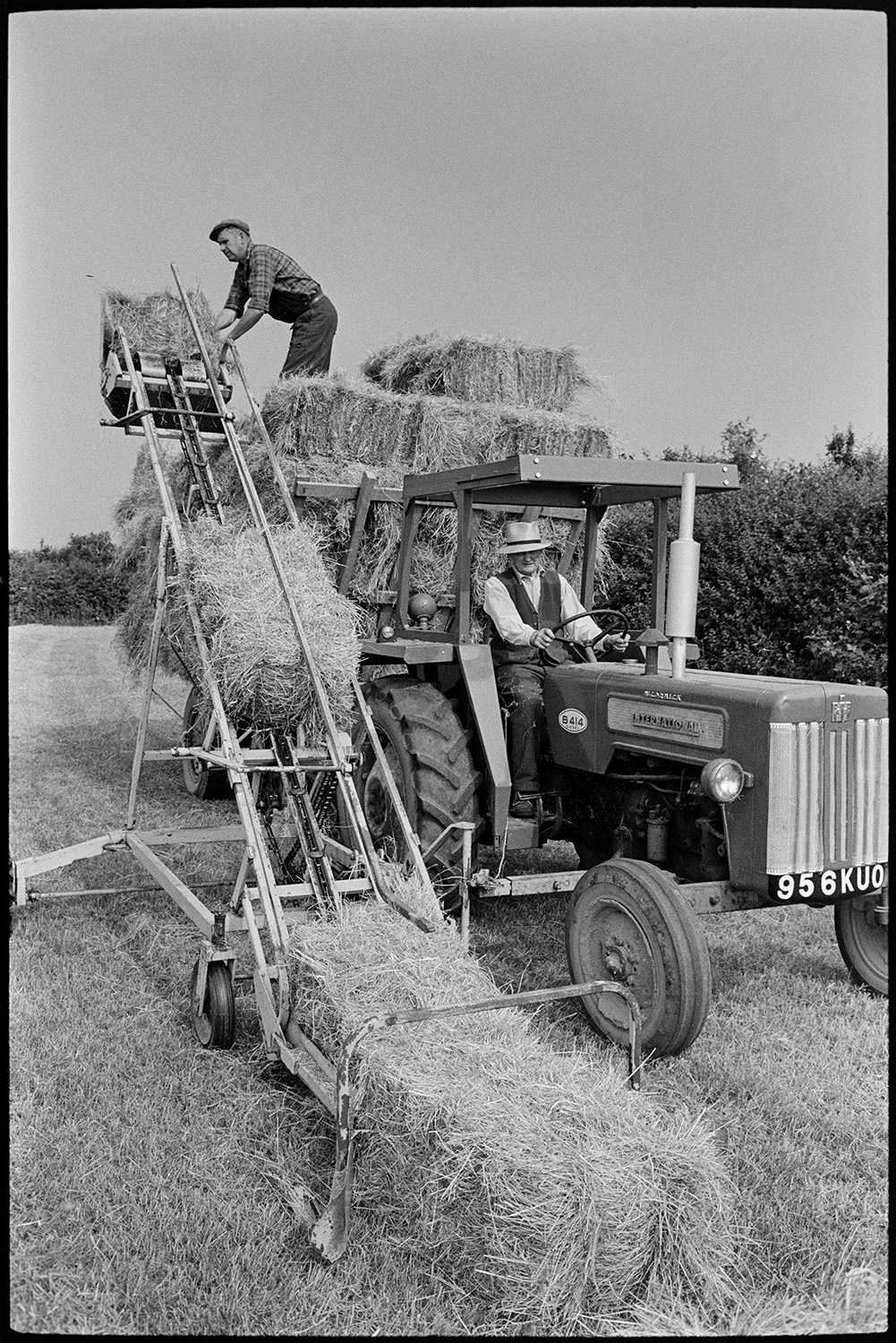 Family haymaking in field. 
[Mr Oke loading hay bales onto a trailer using an elevator in a field at Deckport, Hatherleigh. Another man is driving the tractor attached to the trailer.]