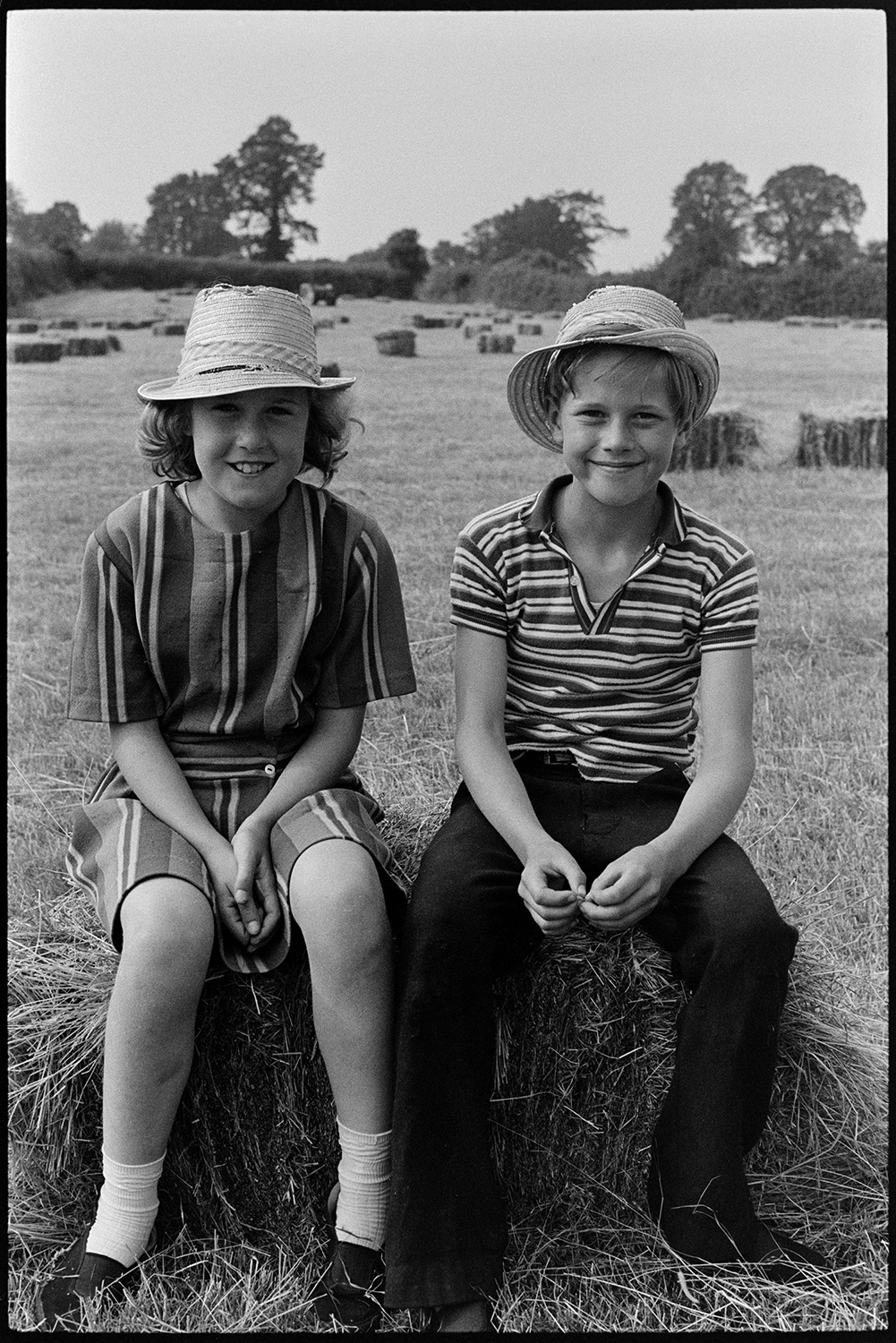 Family haymaking in field. 
[The Oke family twins, a girl and boy, sat on a hay bale in a field at Deckport, Hatherleigh. They are both wearing straw hats. Other hay bales can be seen in the background waiting to be collected.]