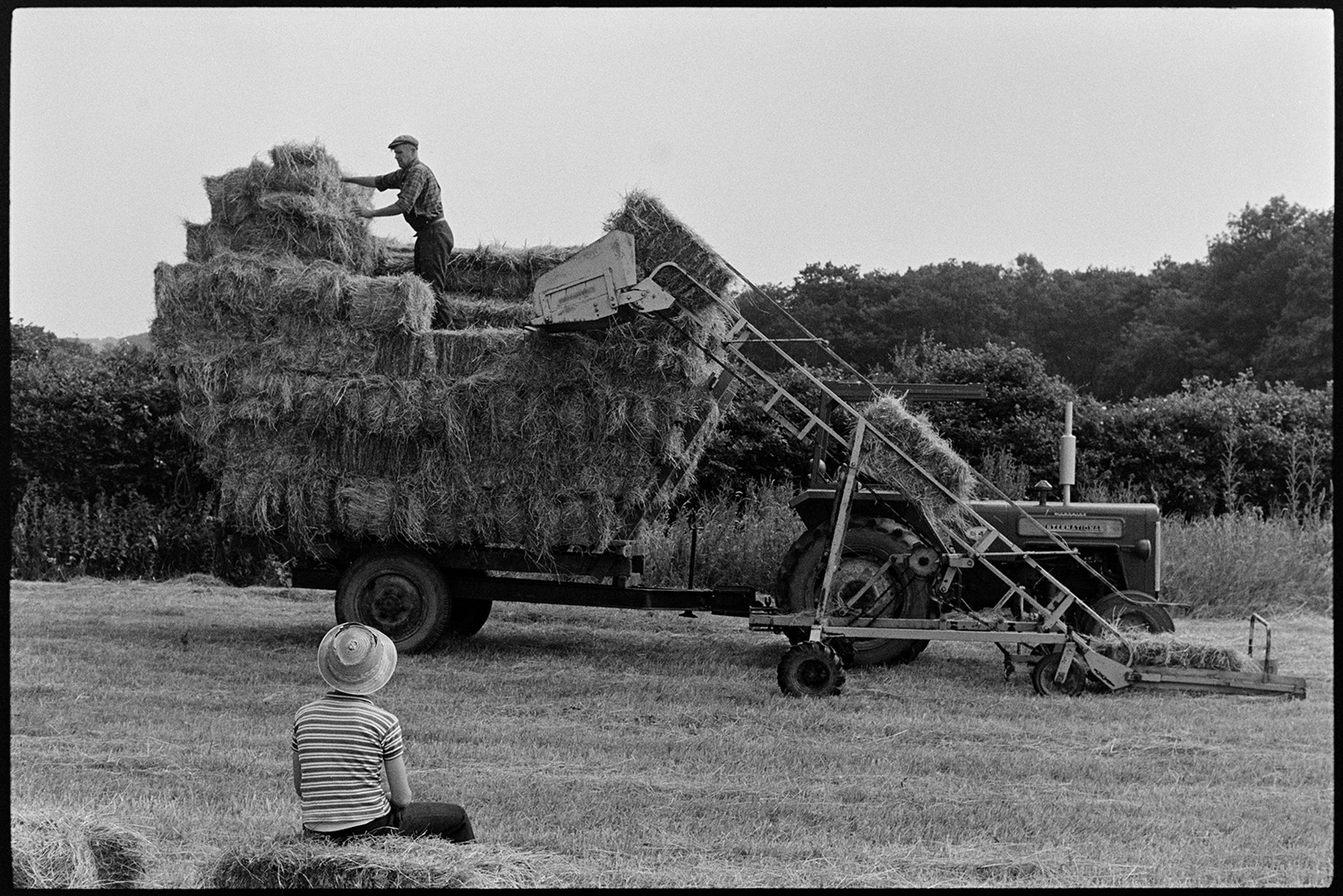 Family haymaking in field. 
[Mr Oke loading hay bales onto a trailer using an elevator in a field at Deckport, Hatherleigh. His son is sat on a hay bale in the field watching him.]