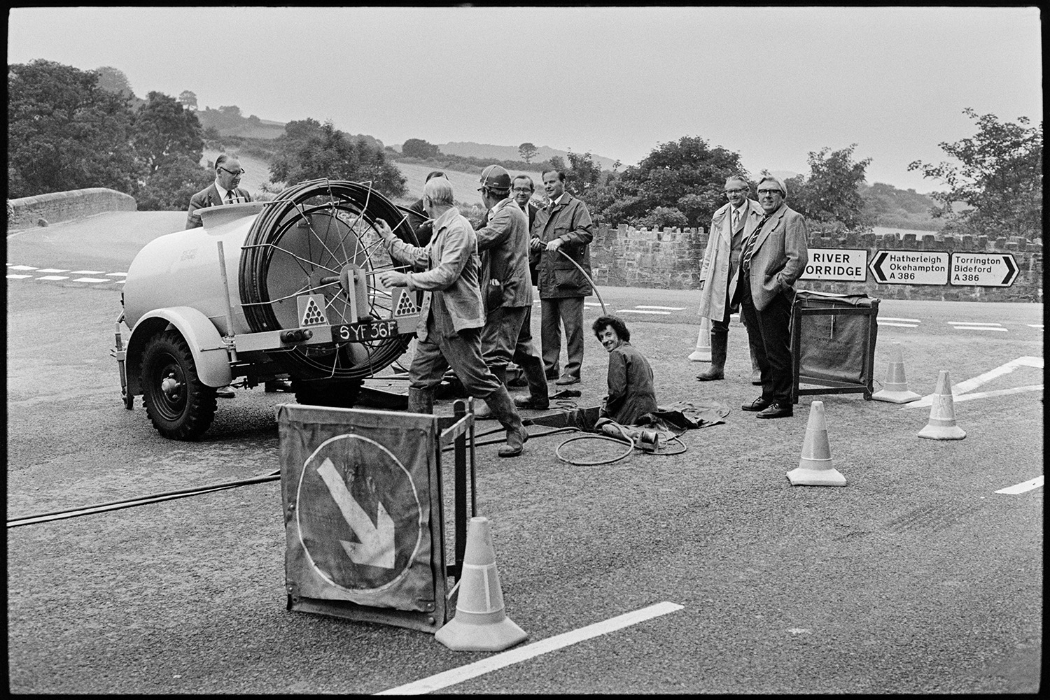 Officials from Electricity Board testing new equipment. 
[Men watching workmen from the Electricity Board test new equipment under a manhole cover in a road by a bridge in Torrington.]
