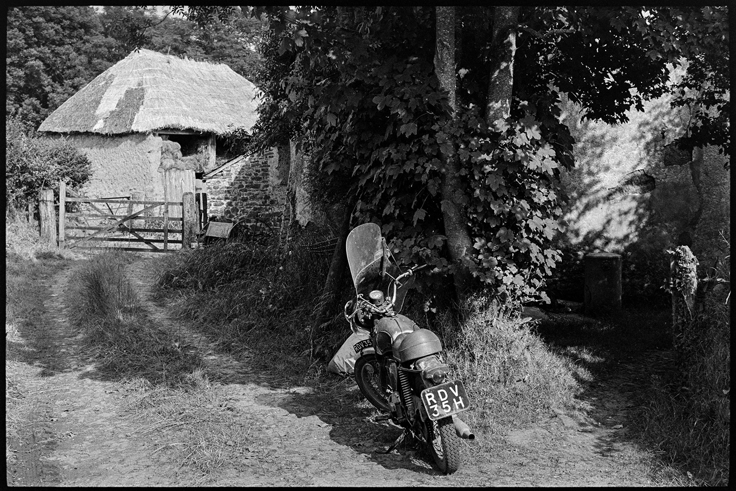Thatcher's motorbike in front of barn. 
[Bill Hammond's motorcycle parked in a lane at Newhouse, Ashreigney. A thatched barn can be seen in the background behind a wooden field gate.]