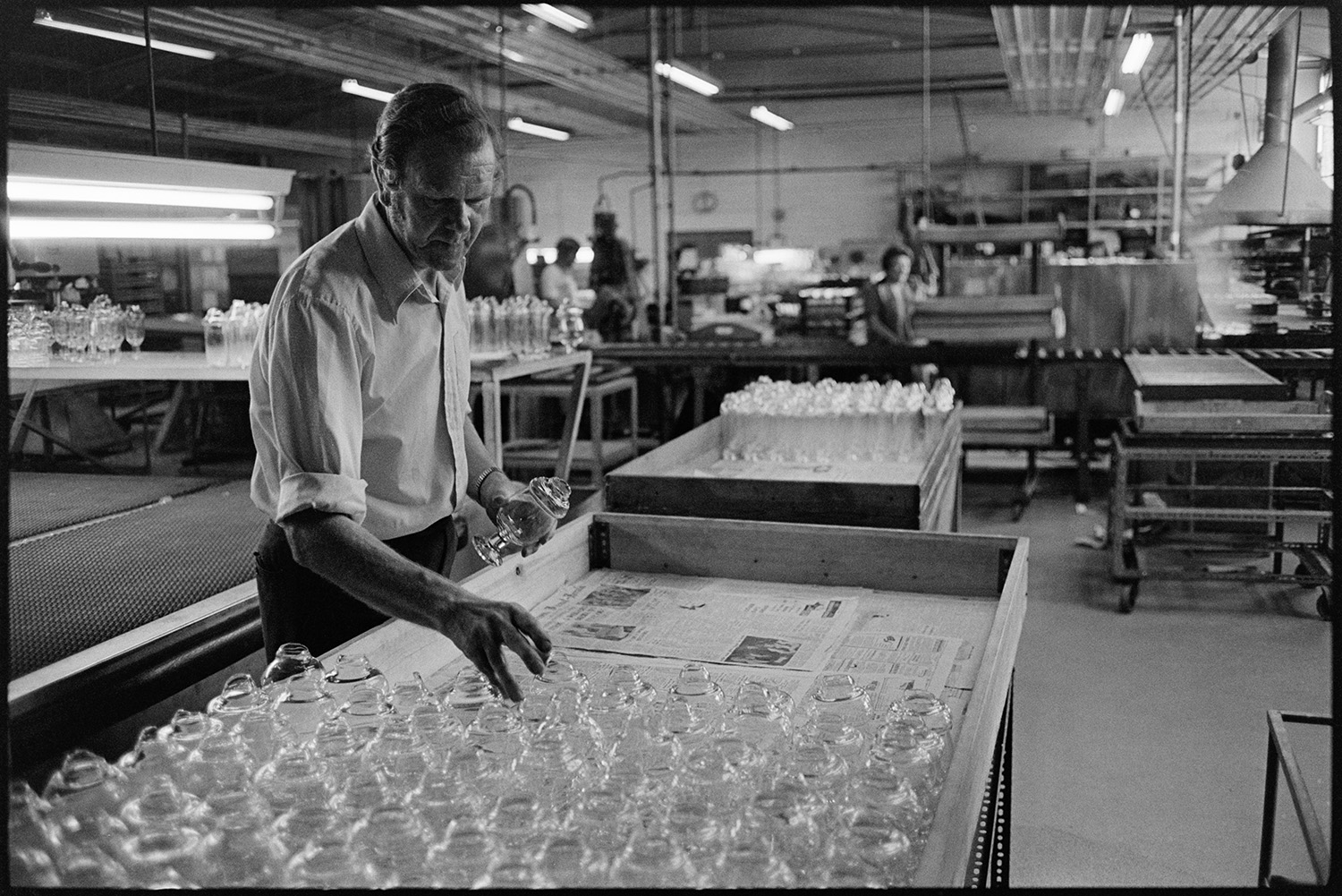 Glass factory with boss. 
[A man checking wine glasses in a wooden tray at a glass factory in Torrington. Various machinery and other trays of glasses can be seen in the background.]