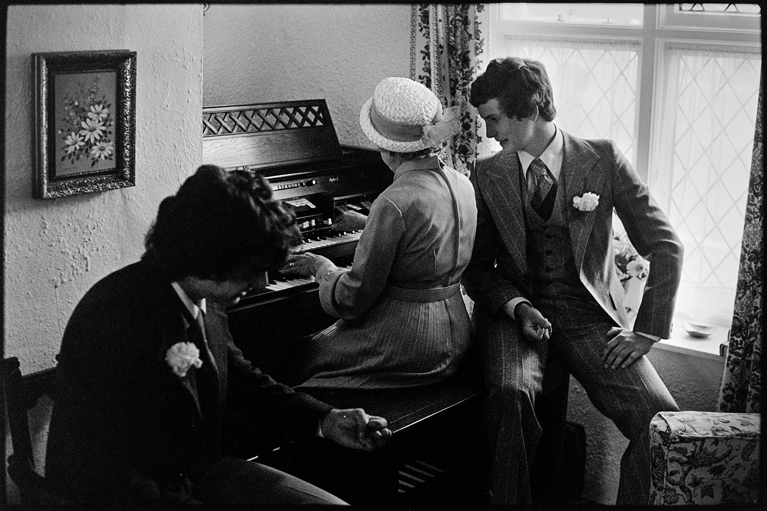 Two men listening to a woman playing an electric organ in a living room. They are wearing buttonholes and have possibly come from or a going to a wedding.