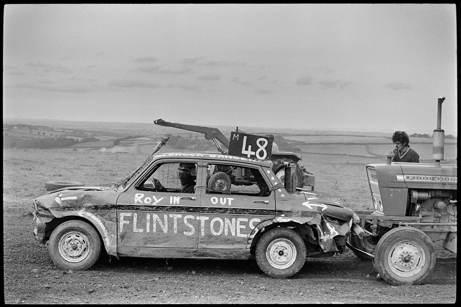 Stock cars at banger races. 
[A driver in a stock car parked in front of a tractor at a banger race at Okehampton. The car has 'Roy In Out Flintstones' written on the side.]