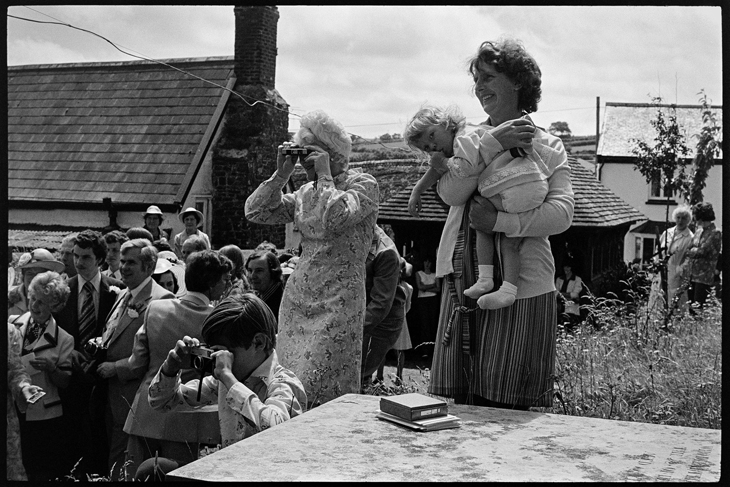 Couple and guests outside church after wedding, photographs, chatting, vicar going home. 
[Wedding guests gathered outside  Atherington Church after Helen Siviter's wedding. A woman and boy are taking photographs while another woman is holding a child.]