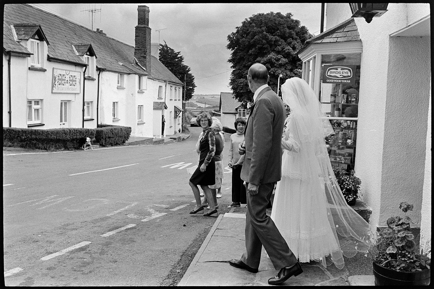 Helen Siviter, the bride, and her father, Geoff Siviter crossing the road to Atherington Church for her wedding. A shop front window is visible behind them and they are talking to passers-by.