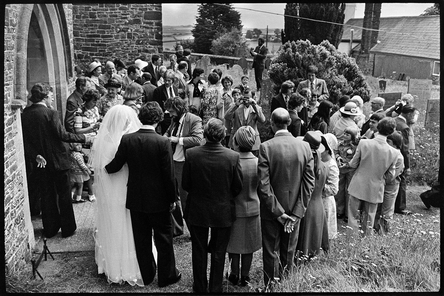 Couple and guests outside church after wedding, photographs, chatting, vicar going home. 
[Helen Siviter, her groom and the bridal party posing for photographs outside Atherington Church after their wedding. Wedding guests are gathered around taking photographs.]