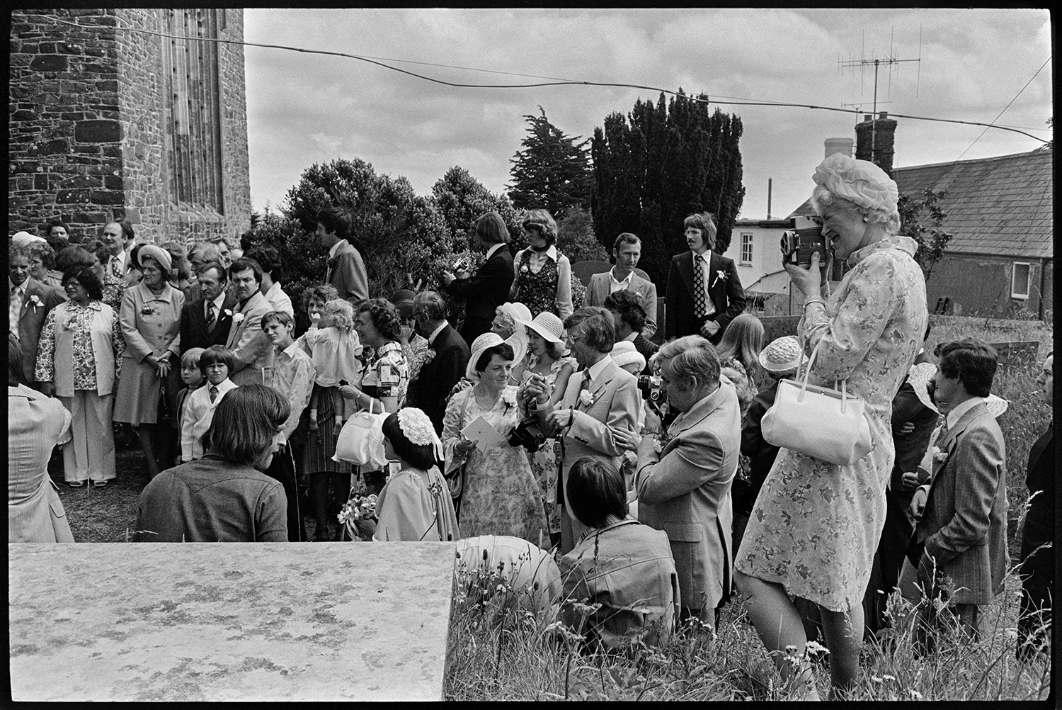 Couple and guests outside church after wedding, photographs, chatting, vicar going home. 
[Wedding guests gathered outside Atherington Church after Helen Siviter's wedding. A woman in the foreground is taking a photograph.]