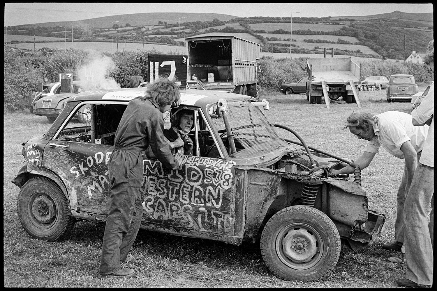Cars, bangers at stock car race. 
[A man talking to a driver in a stock car at a banger race at Okehampton. Another man is looking at the engine of the car. Other parked cars and lorries can be seen in the background.]