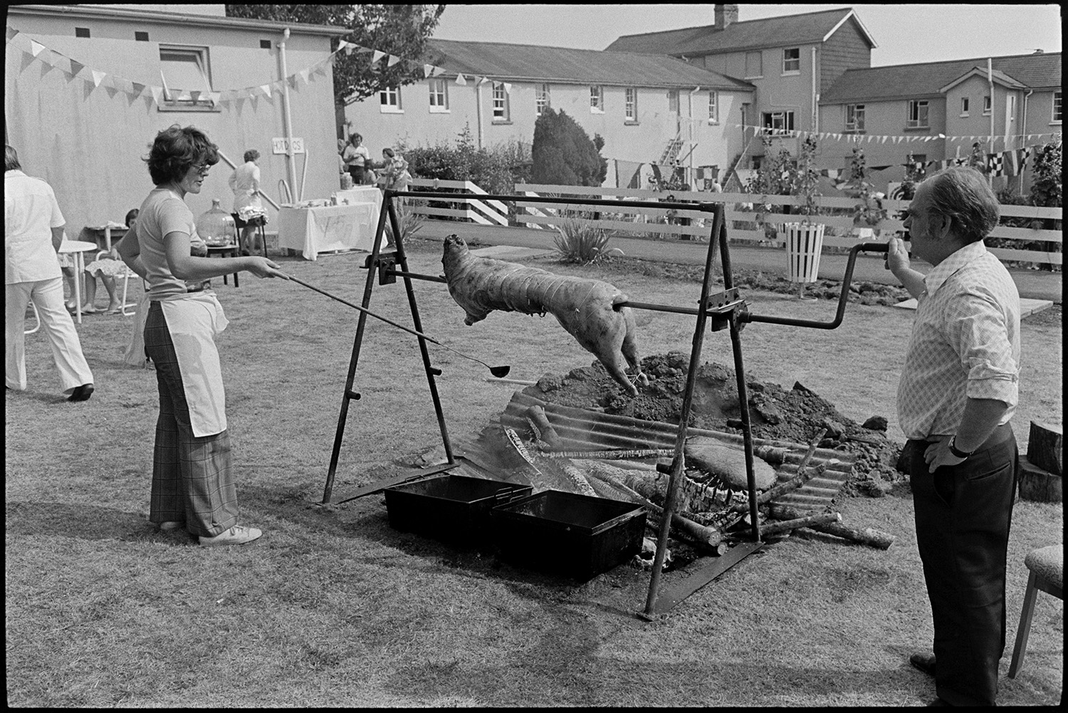 Spit with pig or lamb being barbequed in grounds of sheltered homes. 
[A man and woman spit roasting a pig or lamb at a fete at Torridge View sheltered housing in Torrington. Bunting and stalls can be seen in the background.]